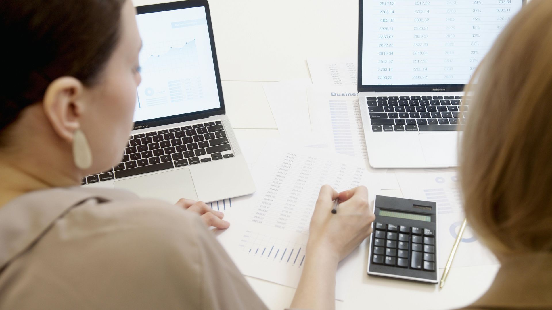 Two people reviewing spreadsheets on laptops with a calculator at a bright office desk